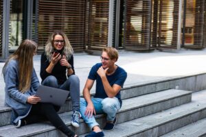 Three students sitting on outdoor steps, chatting and smiling, with a laptop and smartphone.