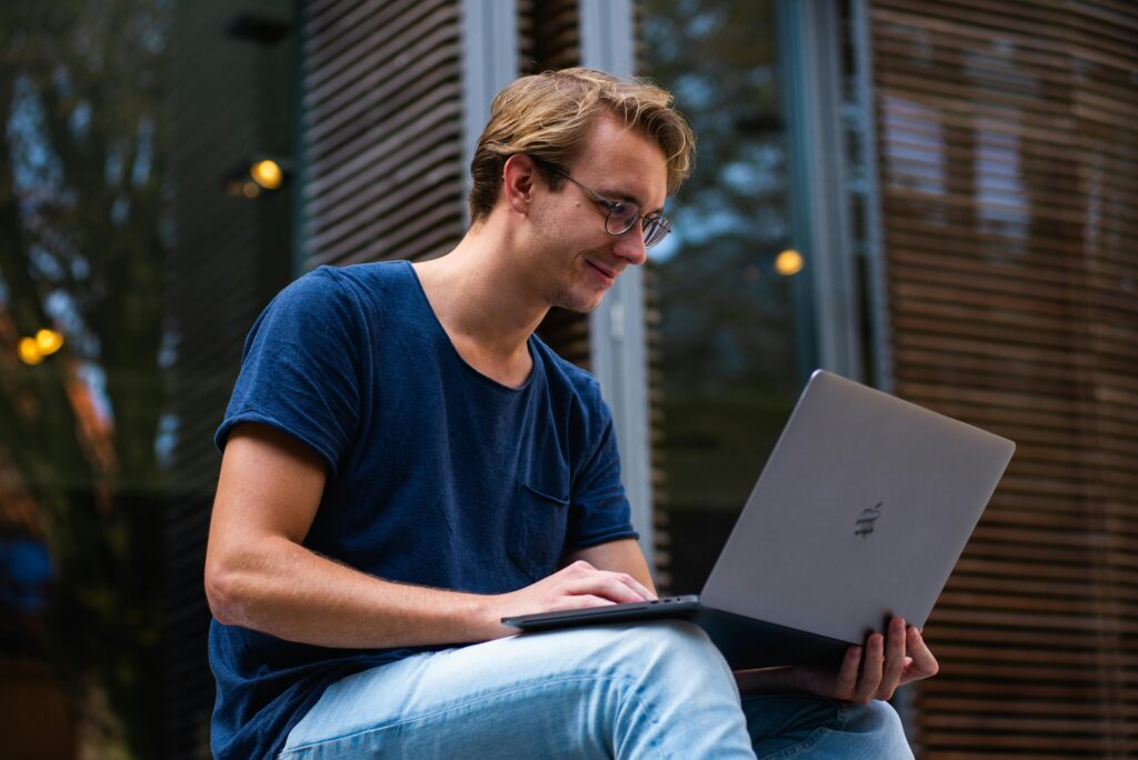 Young man wearing glasses and a blue T-shirt, sitting outdoors with a laptop, smiling while working.